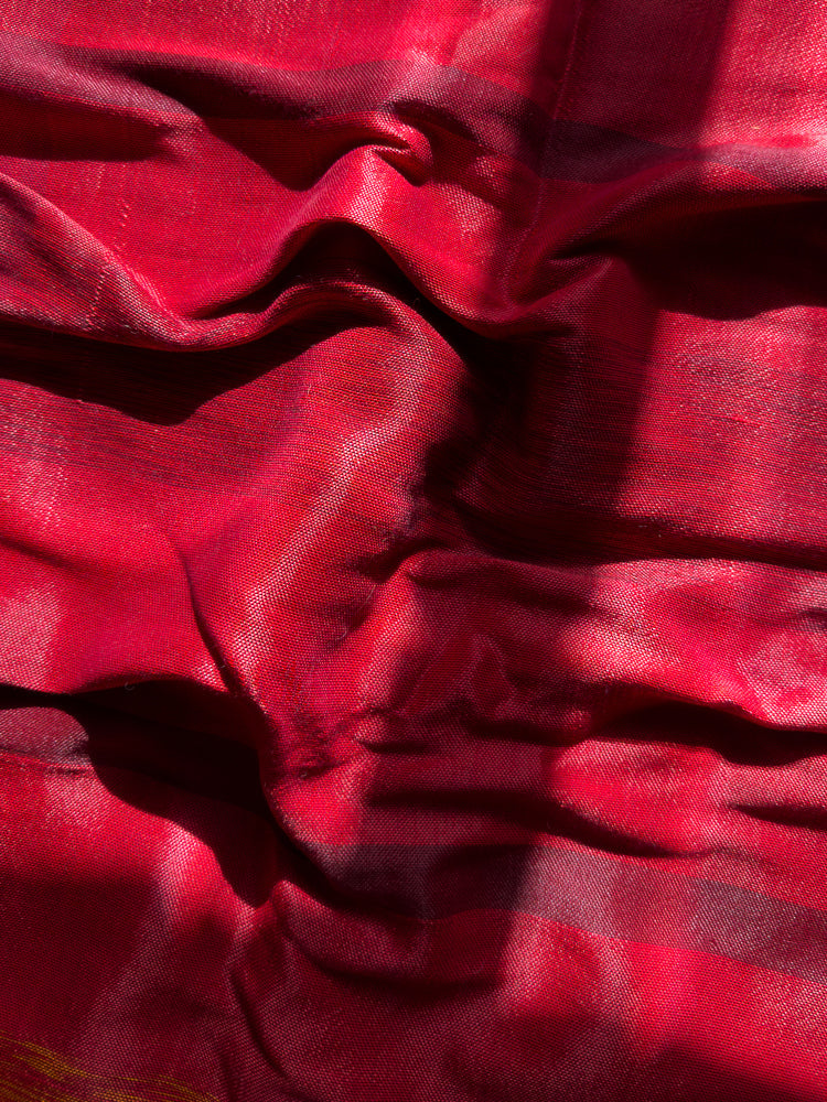 A close-up shot of a red fabric with a textured weave. The fabric is draped and folded, creating shadows and highlights that emphasize its texture and sheen. The lighting suggests it is outdoors or near a window.