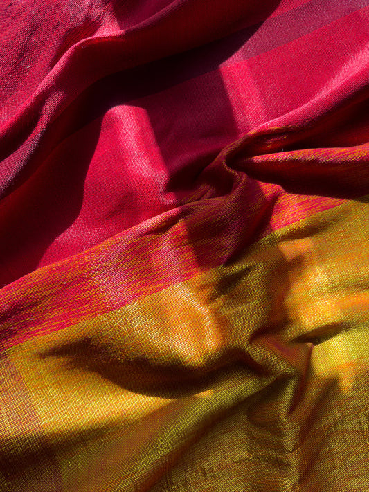 A close-up shot of a richly textured fabric with a gradient of colors from deep red to vibrant yellow and orange. The fabric is draped and folded, creating shadows and highlights that emphasize its weave and sheen.