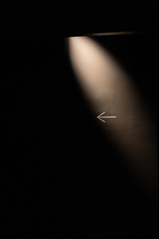 A white arrow points left against a dark background, illuminated by a spotlight from the top left.