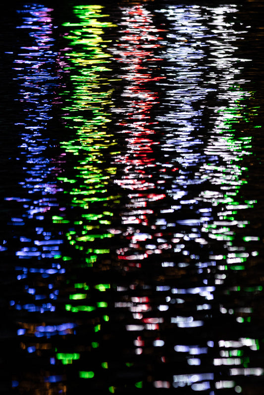 Abstract pattern of colorful lights reflecting on dark water, creating streaks of blue, green, red, and white.