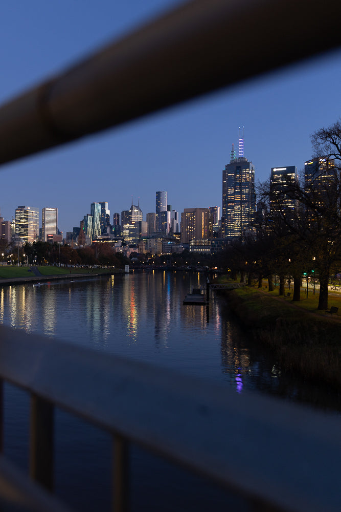 A city skyline at dusk, with illuminated buildings reflected in a calm river. The foreground is blurred by a railing, creating a framed view of the urban landscape.