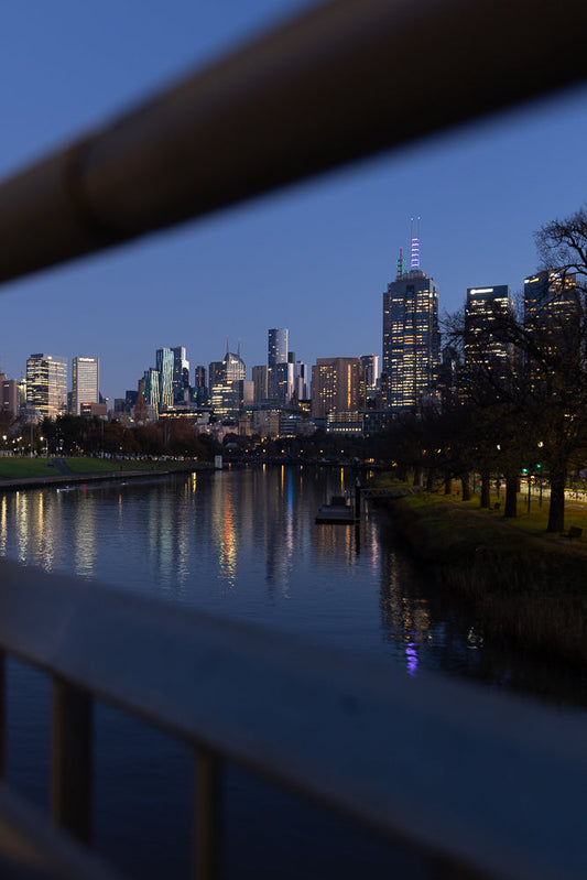 A city skyline at dusk, with illuminated buildings reflected in a calm river. The foreground is blurred by a railing, creating a framed view of the urban landscape.