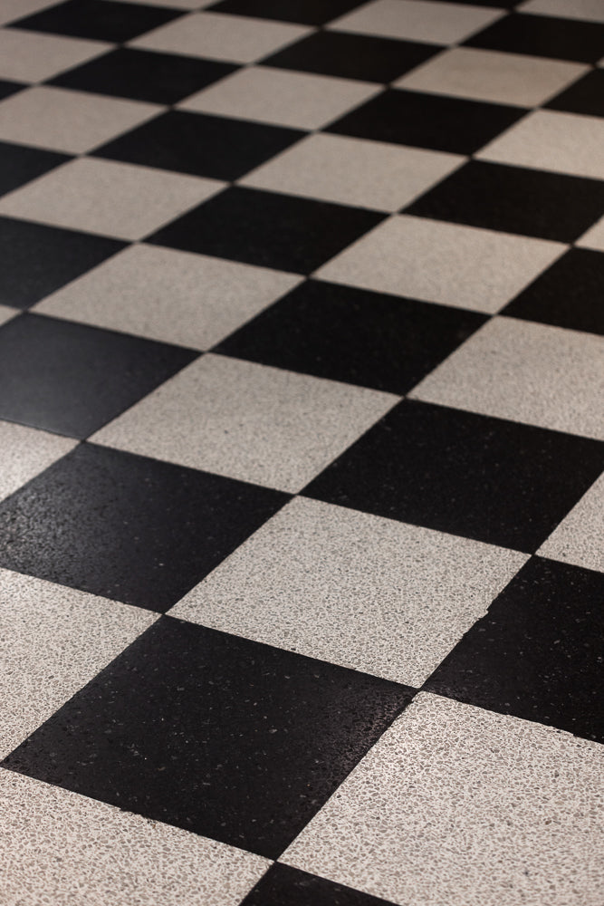 A close-up, angled view of a black and white checkered floor. The tiles are large and square, laid out in a diamond pattern. The lighting creates shadows that emphasize the texture of the tiles.