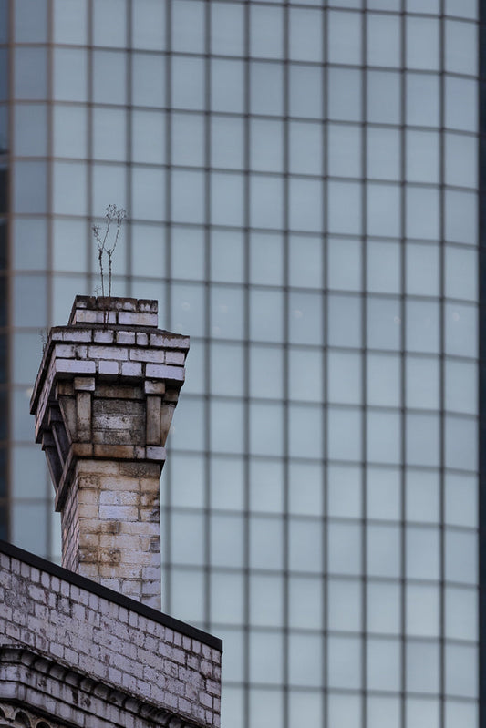 A close-up shot of an old, weathered brick chimney with a few dry plants growing from the top. In the background, a modern glass building with a grid pattern is visible.