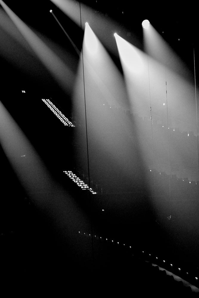 A black and white image of stage lights creating beams of light in a dark venue. Several rows of lights are visible, some are bright spotlights, and others are smaller, more numerous lights arranged in rows.
