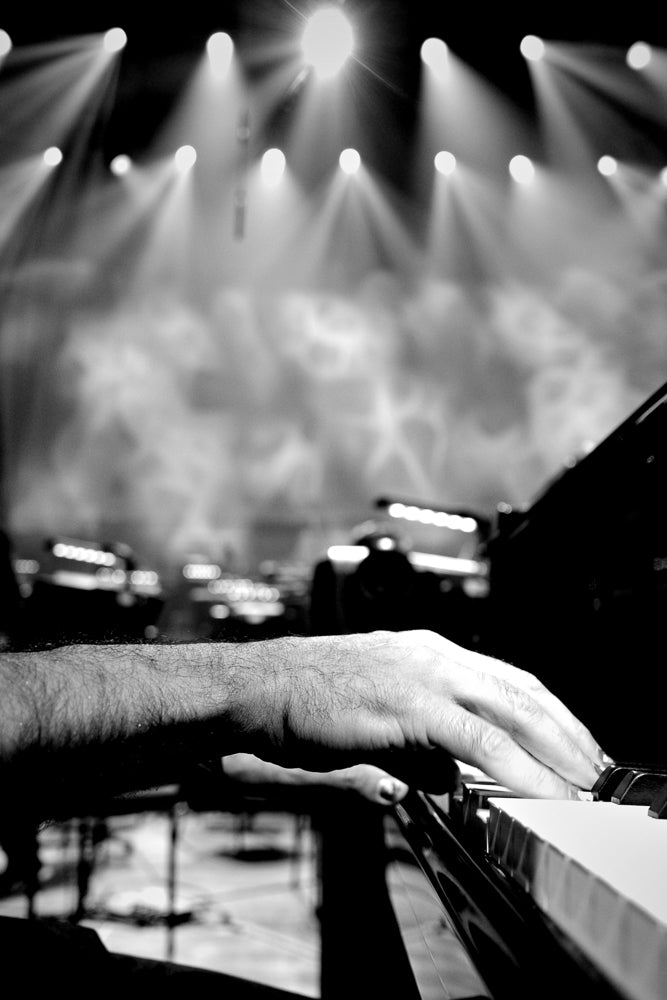 A close-up, black and white shot shows a pianist's hands on the keys of a grand piano. The background is blurred, with stage lights and smoke creating a dramatic atmosphere.