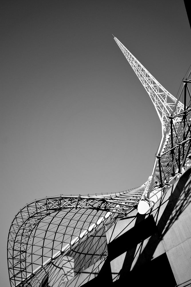A black and white, low-angle shot of the Melbourne Cricket Ground's stadium roof structure against a clear sky. The intricate metal framework of the roof curves and extends upwards, culminating in a sharp, pointed spire. Shadows from the structure are cast across the concrete facade.