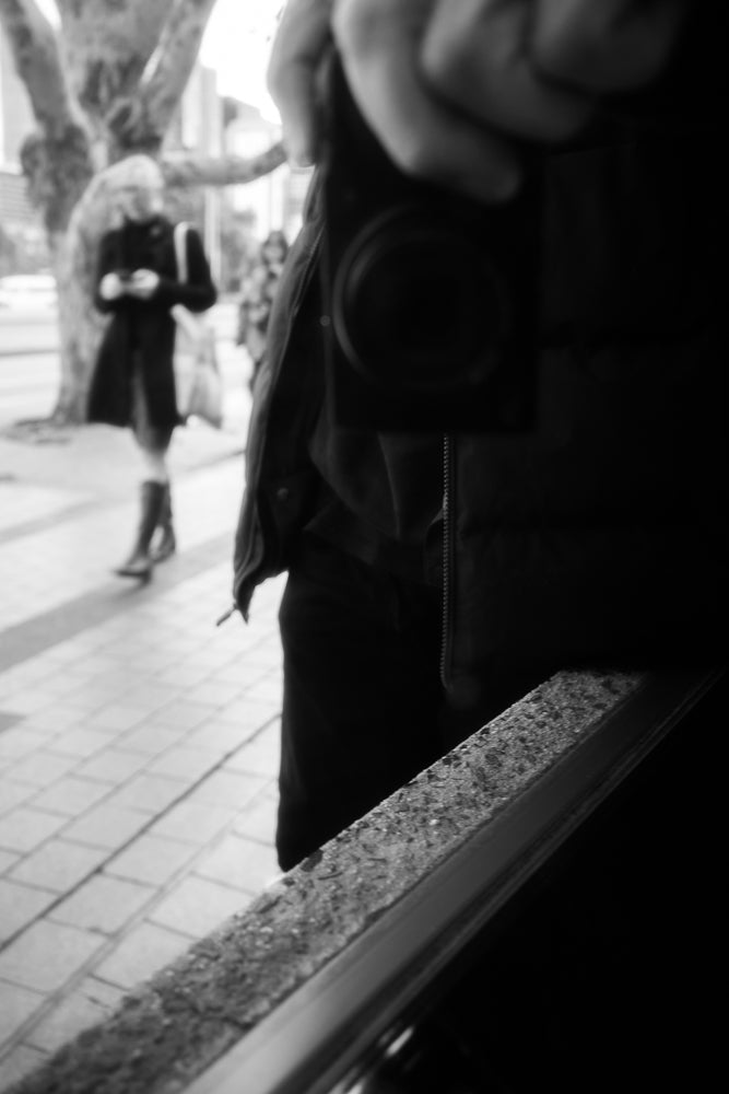A black and white photo shows a person holding a camera, possibly taking a selfie, with another person in the background walking away.