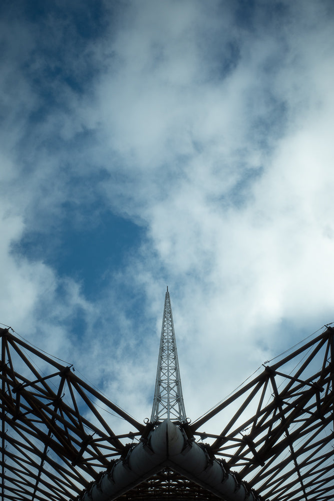 A low-angle shot of a tall, slender radio tower piercing a cloudy blue sky. The tower is framed by the dark, angular supports of a large structure, creating a dramatic and geometric composition.