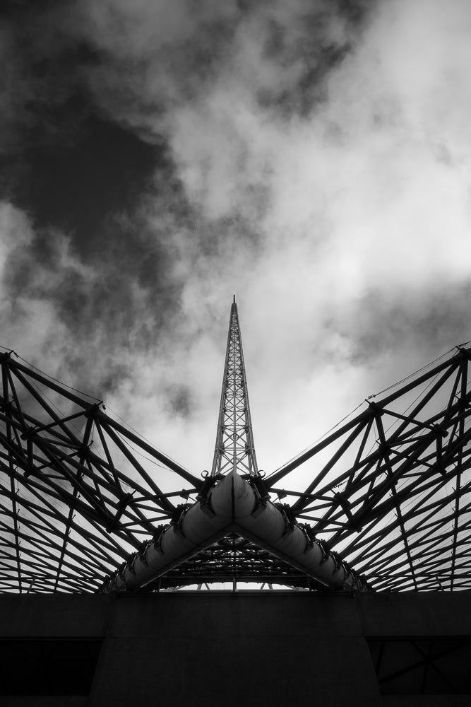 A dramatic, low-angle, black and white shot of a tall, lattice-like tower rising into a cloudy sky, framed by the geometric structure of a stadium roof.