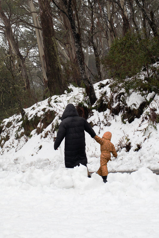 A person in a black puffer coat holds hands with a child in a tan snowsuit as they walk through deep snow in a forest with tall, thin trees.