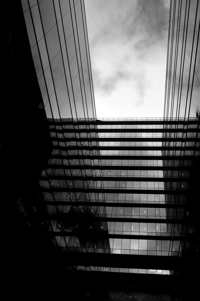 A black and white, low-angle shot of a modern glass building. The building's facade is made of repeating horizontal bands of windows, separated by dark, horizontal beams. The sky above is cloudy and bright, contrasting with the dark, reflective glass of the building.