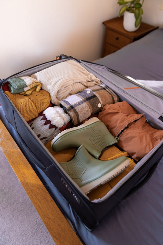 A suitcase is packed with warm clothing for a trip. Items include a cream sweater, a plaid shirt, a brown jacket, yellow and green mittens, and two pairs of green rain boots.