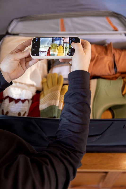 A person is taking a photo of a suitcase packed with winter clothes, including sweaters, gloves, and boots.