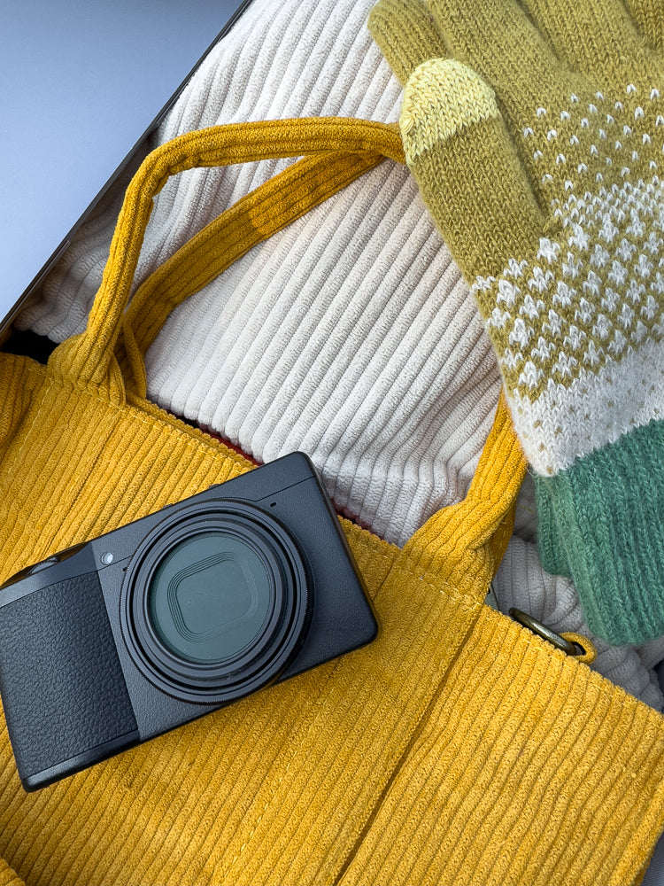 A black digital camera rests on a yellow corduroy tote bag, next to a pair of green and yellow knit gloves and a white ribbed sweater.