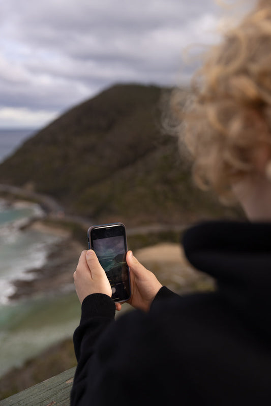 A person with curly blonde hair, wearing a black hoodie, holds a smartphone in their hands, capturing a scenic view of a coastline with waves crashing on the shore and a winding road along a hill.