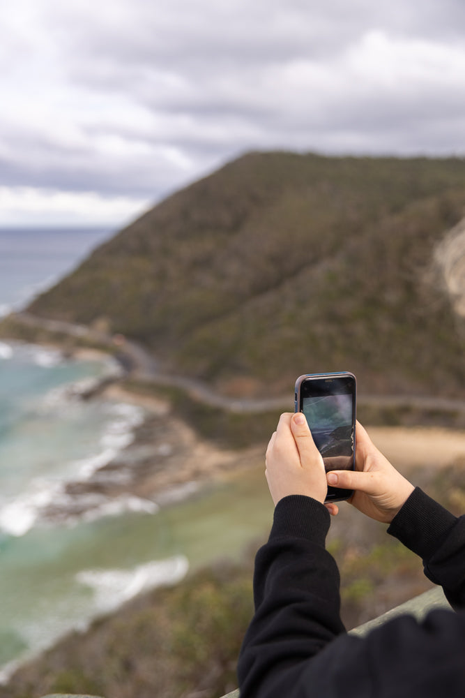 A person holds a smartphone to take a photo of a scenic coastal road winding along a rugged hillside overlooking the ocean under a cloudy sky.