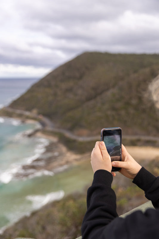 A person holds a smartphone to take a photo of a scenic coastal road winding along a rugged hillside overlooking the ocean under a cloudy sky.