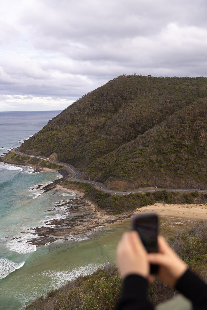 A person holds a smartphone to take a photo of a winding coastal road with the ocean and a forested hill. The sky is overcast with clouds.