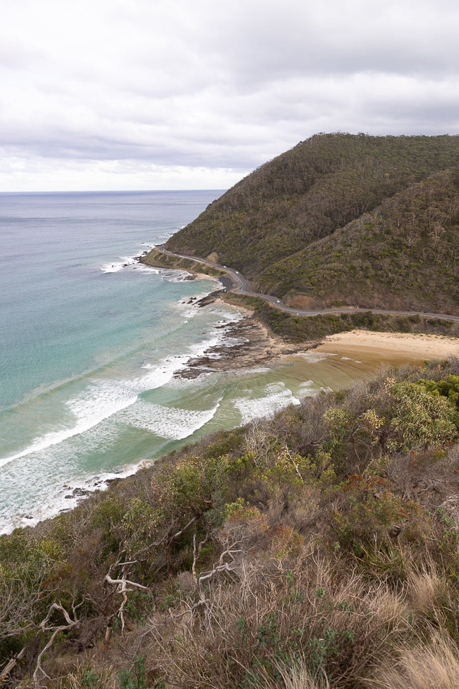 A winding road hugs the coastline, with the turquoise ocean on one side and a densely forested hill on the other. Waves crash onto a sandy beach below the road.