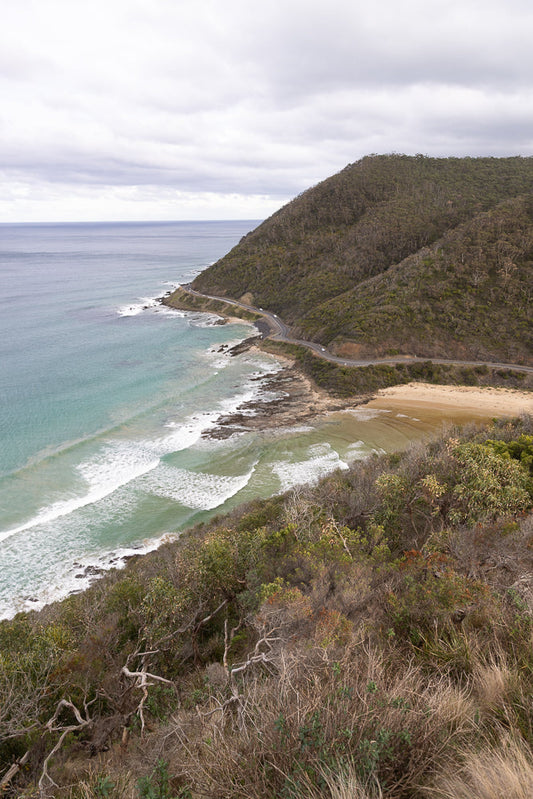 A winding road hugs the coastline, with the turquoise ocean on one side and a densely forested hill on the other. Waves crash onto a sandy beach below the road.