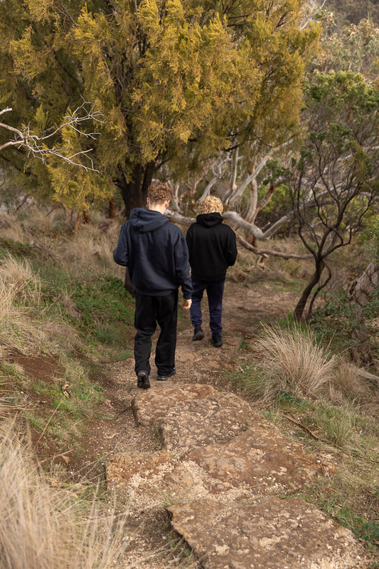 Two young people walk away from the camera on a rocky path through a forest with yellow and green foliage.