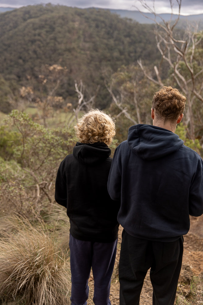 Two young people, viewed from behind, stand on a dirt path looking out over a forested valley under a cloudy sky. The person on the left has curly blonde hair and wears a black hoodie and blue pants. The person on the right has curly brown hair and wears a dark blue hoodie and black pants.