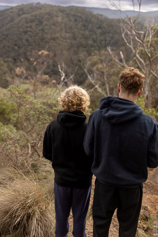 Two young people, viewed from behind, stand on a dirt path looking out over a forested valley under a cloudy sky. The person on the left has curly blonde hair and wears a black hoodie and blue pants. The person on the right has curly brown hair and wears a dark blue hoodie and black pants.