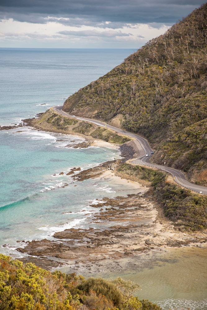 A winding road hugs the coastline of a rugged, tree-covered hill overlooking the ocean. The turquoise water crashes against rocky shores, with a white car visible on the road.