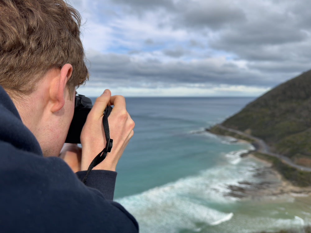A person in a dark blue hoodie holds a camera to their face, taking a photo of a scenic coastal view with turquoise water, white waves, and a winding road along a green hillside under a cloudy sky.