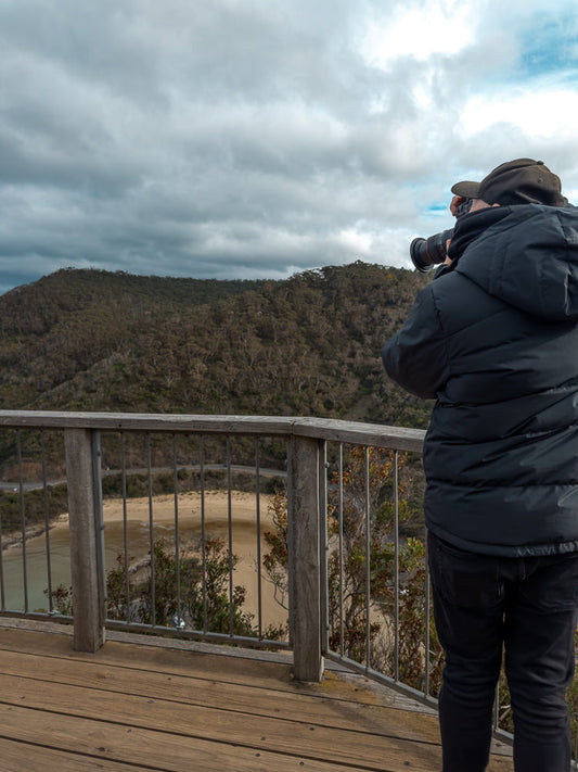 A person wearing a black puffer jacket and a baseball cap stands on a wooden platform, taking a photo with a large camera. They are looking out at a scenic view of a forested hill and a beach with a river.