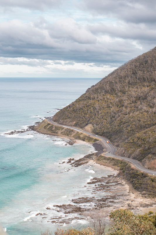 A winding road hugs the coastline of a rugged, tree-covered mountain, with the turquoise ocean stretching out to the horizon under a cloudy sky. A few cars are visible on the road, suggesting travel and exploration along this scenic route.