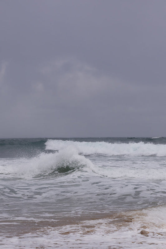 A grey, overcast sky hangs over a choppy ocean with several waves breaking. White foam and spray are visible as the waves crash onto the shore.