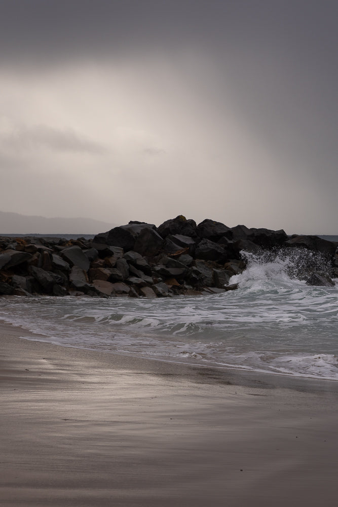 A wave crashes against a rocky breakwater on a cloudy day. The wet sand on the beach reflects the overcast sky.