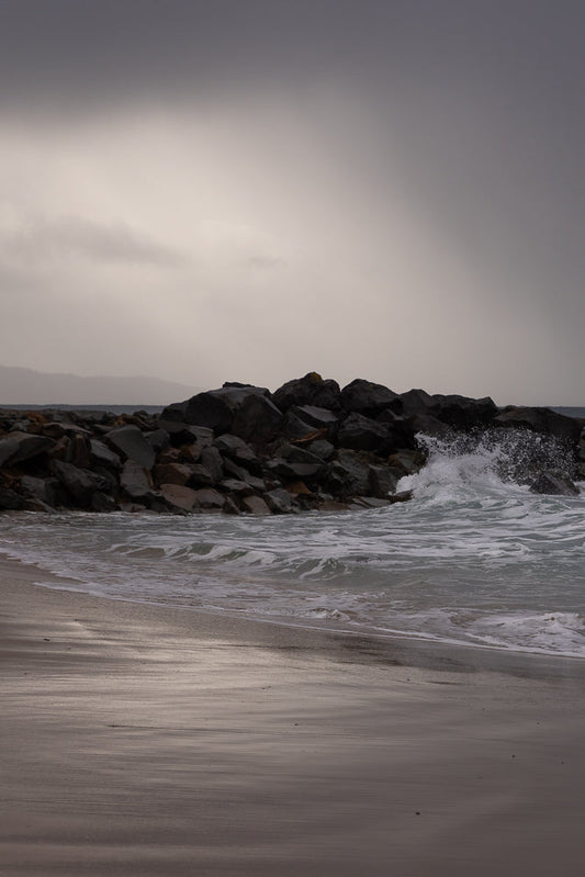 A wave crashes against a rocky breakwater on a cloudy day. The wet sand on the beach reflects the overcast sky.