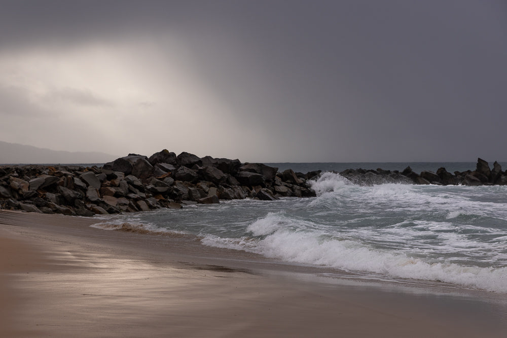 A rocky breakwater juts into the ocean under a stormy, overcast sky. Waves crash against the rocks and wash onto a sandy beach in the foreground.