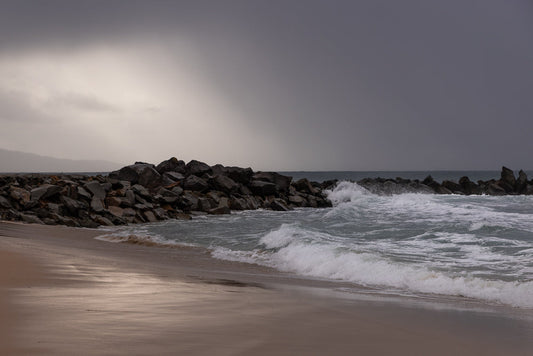 A rocky breakwater juts into the ocean under a stormy, overcast sky. Waves crash against the rocks and wash onto a sandy beach in the foreground.