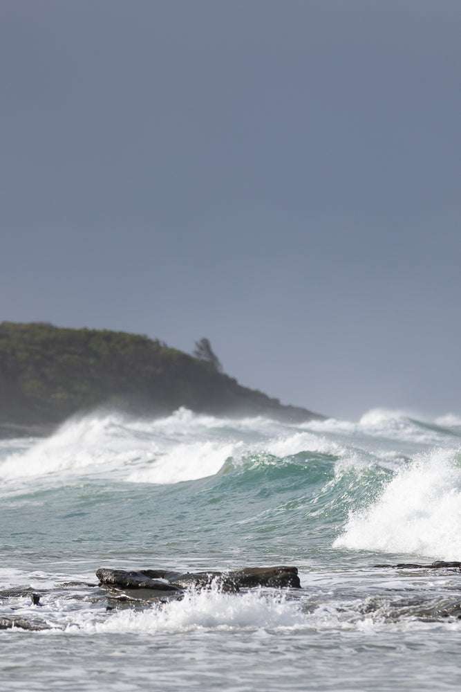 Waves crash against rocks on a beach with a tree-covered hill in the background under a cloudy sky.