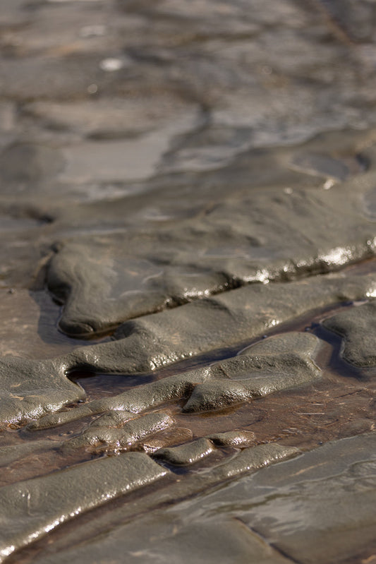 Close-up of wet, textured sand and rock formations with shallow pools of water reflecting sunlight. The patterns create a natural, abstract landscape.