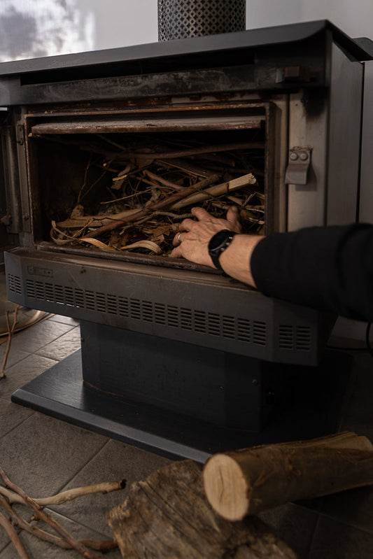 A person's hand with a watch is reaching into a wood-burning stove filled with branches and leaves.