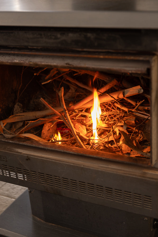 A close-up shot of a fireplace with dry twigs and leaves burning. Flames are visible, casting a warm glow on the surrounding materials.