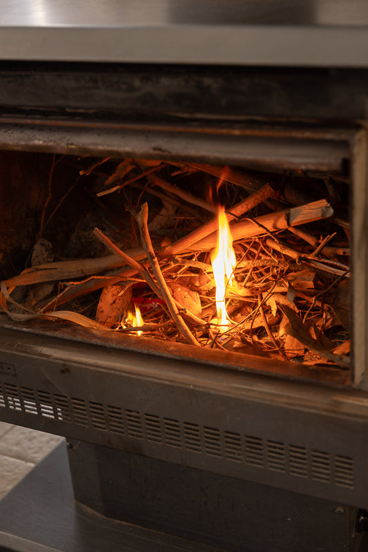 A close-up shot of a fireplace with dry twigs and leaves burning. Flames are visible, casting a warm glow on the surrounding materials.