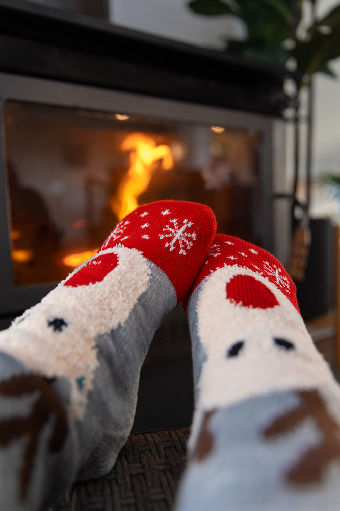 A close-up shot of a person's feet wearing festive reindeer socks, resting in front of a warm, glowing fireplace. The socks are red with white snowflakes and feature reindeer faces.