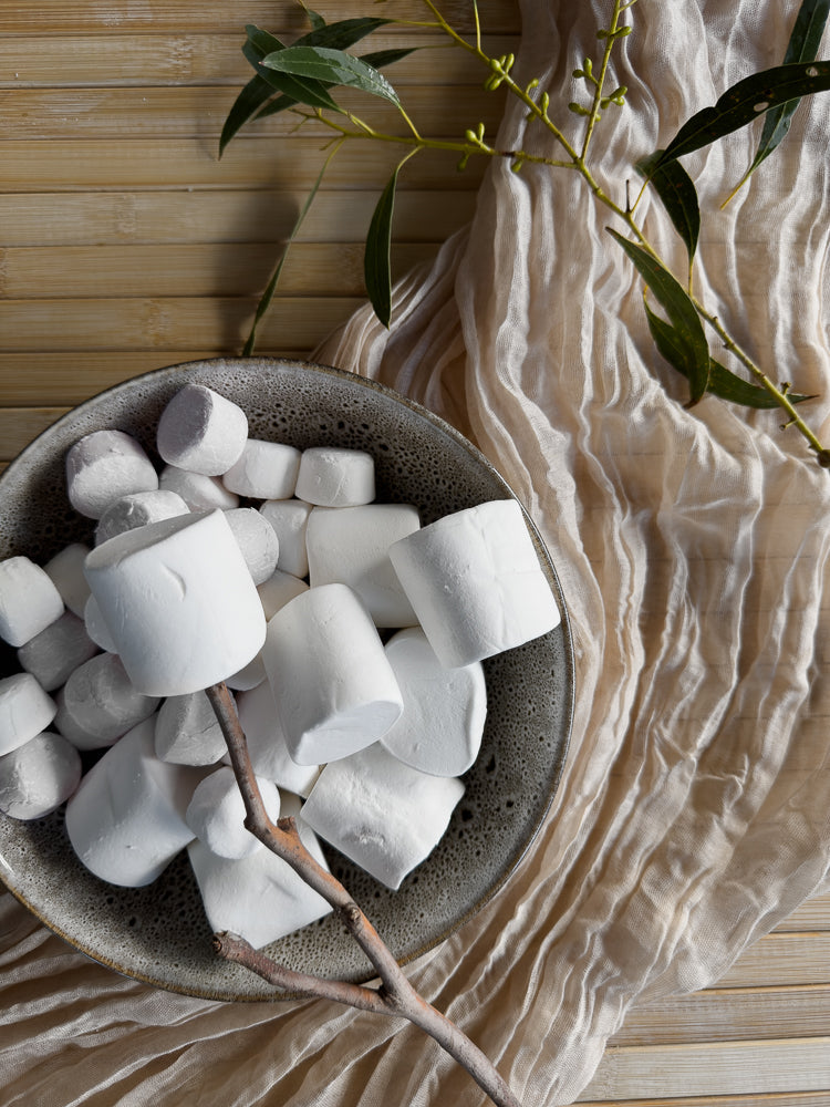 A bowl of white marshmallows sits on a textured beige fabric, with a eucalyptus branch draped over the scene. The marshmallows are soft and fluffy, some are on a stick.