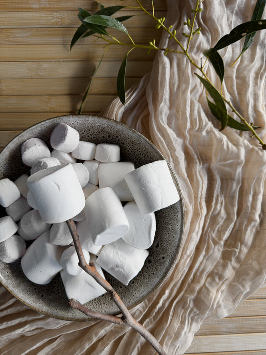 A bowl of white marshmallows sits on a textured beige fabric, with a eucalyptus branch draped over the scene. The marshmallows are soft and fluffy, some are on a stick.