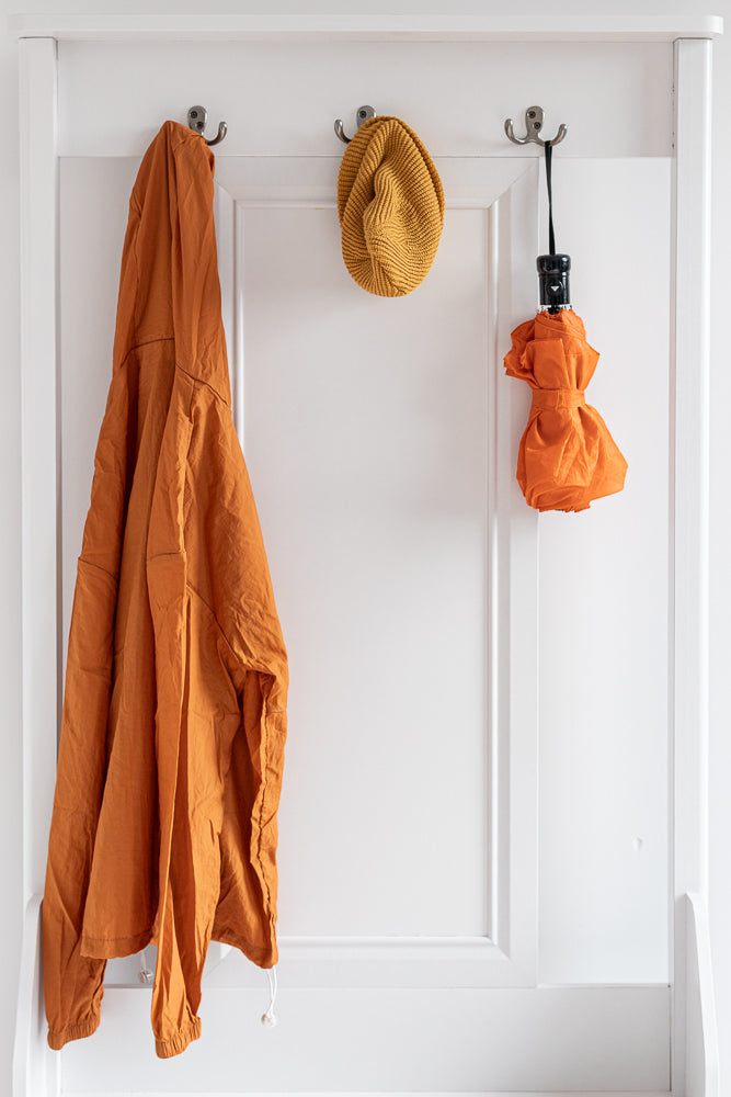 An orange jacket, a yellow knitted hat, and a folded orange umbrella hang on hooks on a white wall.