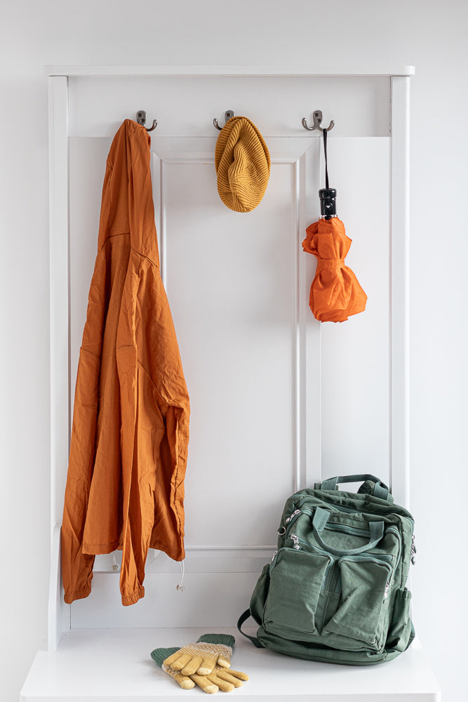 A white coat rack holds an orange jacket, a yellow beanie, and a folded orange umbrella. A green backpack and a pair of yellow and green gloves sit on the shelf below.