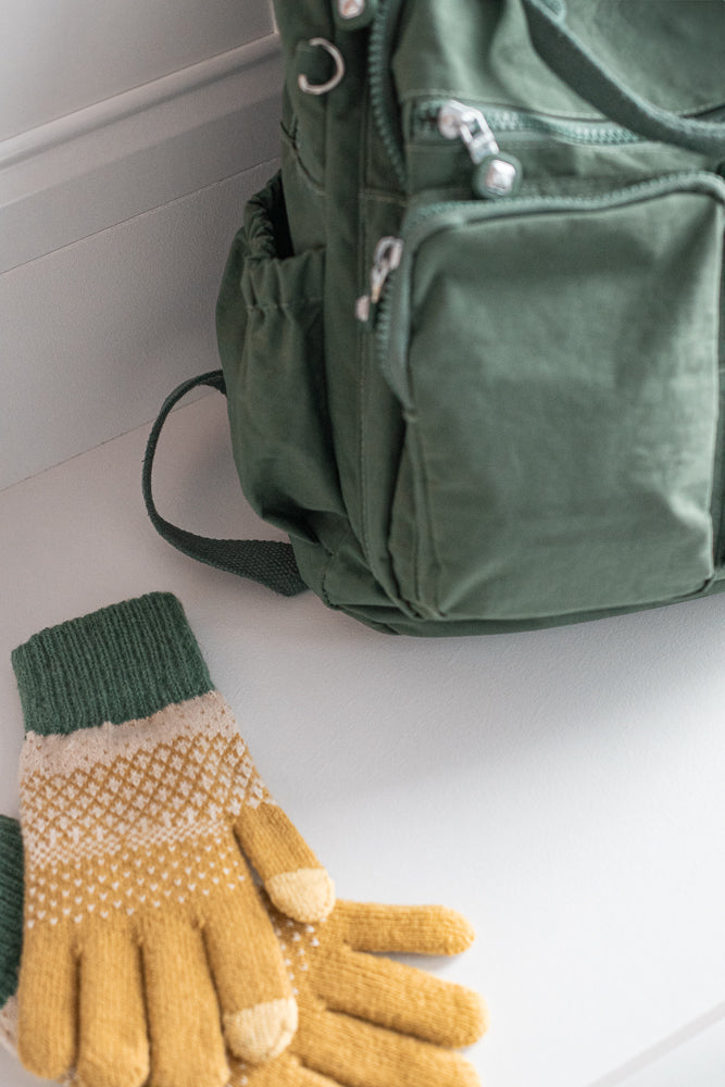 A pair of yellow and white patterned gloves with green cuffs lies on a white surface next to a green backpack.