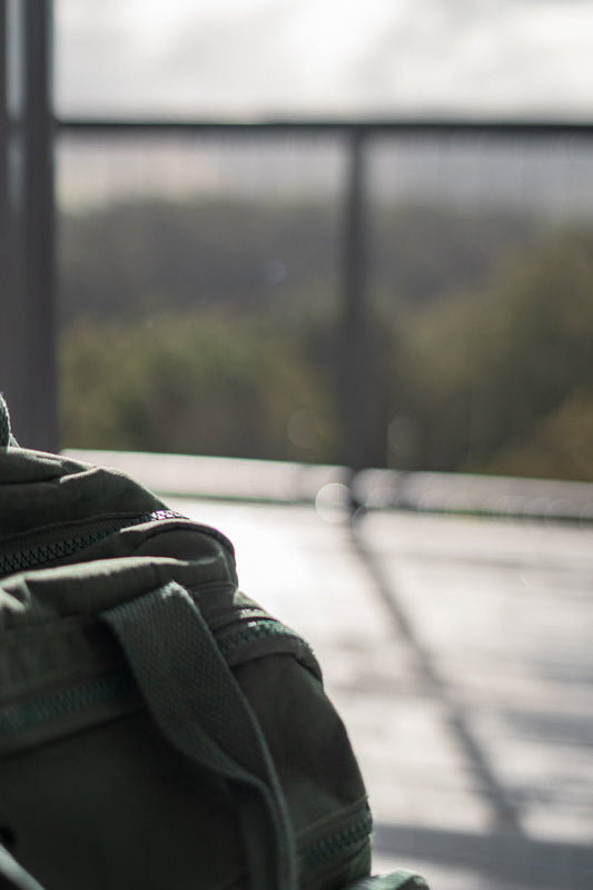 A close-up shot of a green canvas duffel bag with a zipper and strap visible. The background is blurred, showing a hint of trees and a railing.