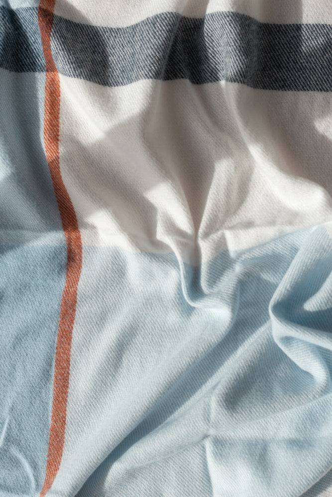 A close-up shot of a soft, textured fabric with a plaid pattern. The fabric features horizontal stripes in white, dark blue, and light blue, with a vertical orange stripe running through the center.